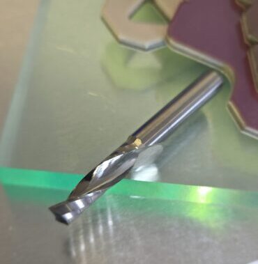 Close-up of a shiny twist drill bit resting on a metal work surface with blurred colorful shapes in the background.