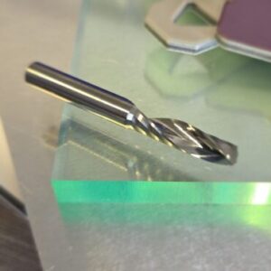 Close-up of a silver twist drill bit resting on a metallic surface with blurred colorful shapes in the background (industrial/working scene).