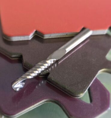 Close-up of a metal twist drill bit resting on layered purple and black metal pieces with a blurred red card in the background.