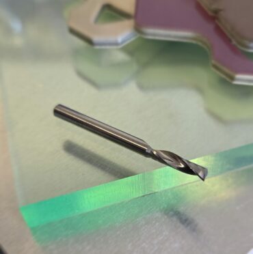 Close-up of a metal twist drill bit resting on a reflective glass surface with blurred colorful shapes in the background.