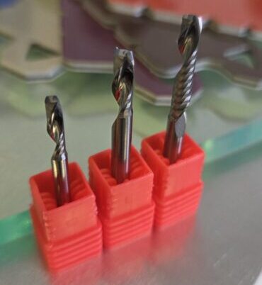 Three metal twist drill bits held in red plastic blocks on a stainless worktable, with a colorful, blurred background.