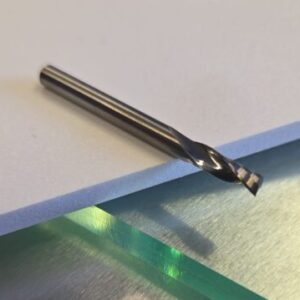 Close-up of a metallic drill bit lying on a white foam board atop a brushed metal work surface, with a blurred circular glass object in the background.