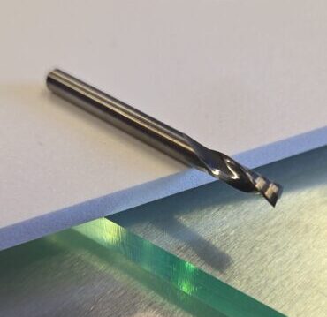 Close-up of a metallic drill bit lying on a white foam board atop a brushed metal work surface, with a blurred circular glass object in the background.