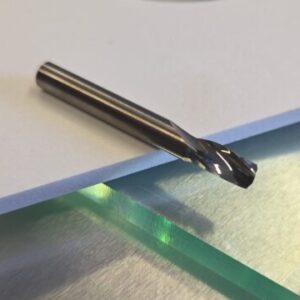 Close-up of a metallic drill bit lying on a white foam sheet with a blurred circular green-tinted disc in the background.