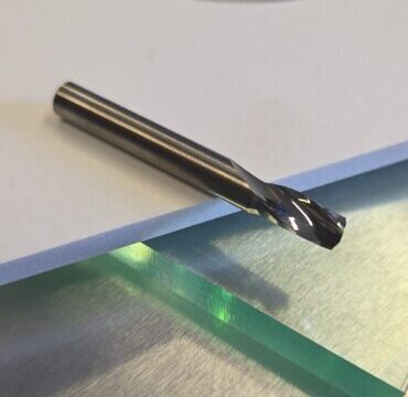 Close-up of a metallic drill bit lying on a white foam sheet with a blurred circular green-tinted disc in the background.